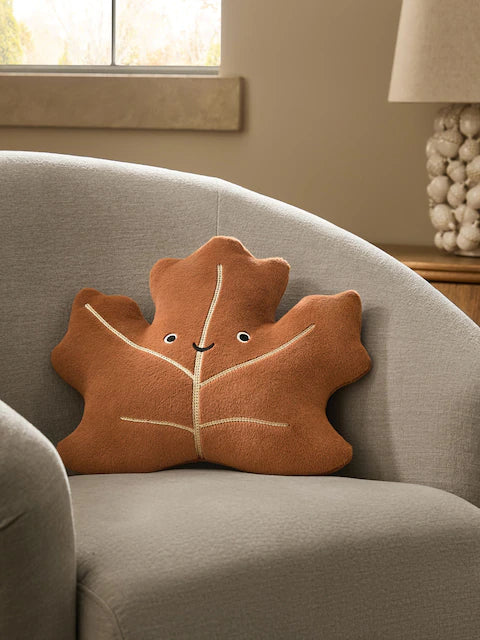3D maple leaf velvet cushion with embroidered smiley face on grey armchair in warm autumn living room