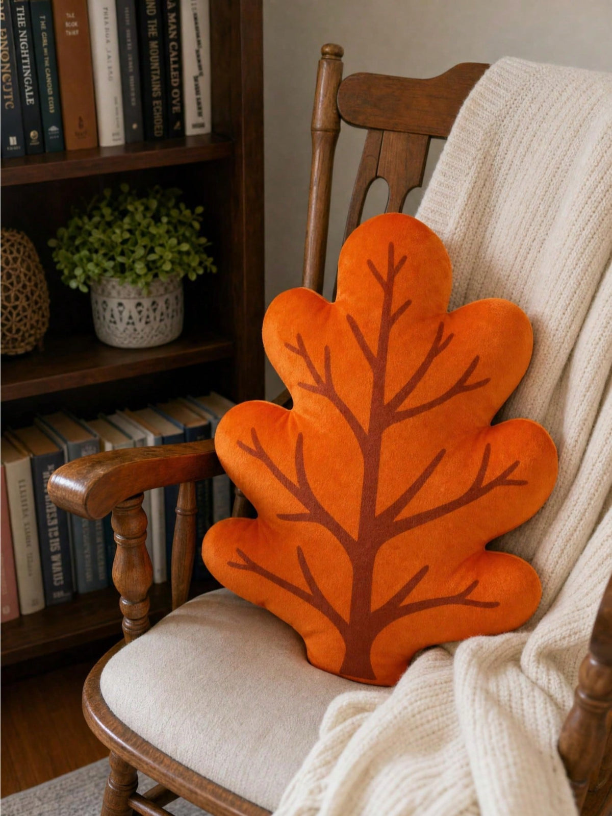 3D oak leaf shaped cushion in burnt orange with dark brown vein detail on vintage wooden rocking chair with cream knit throw and bookshelf background