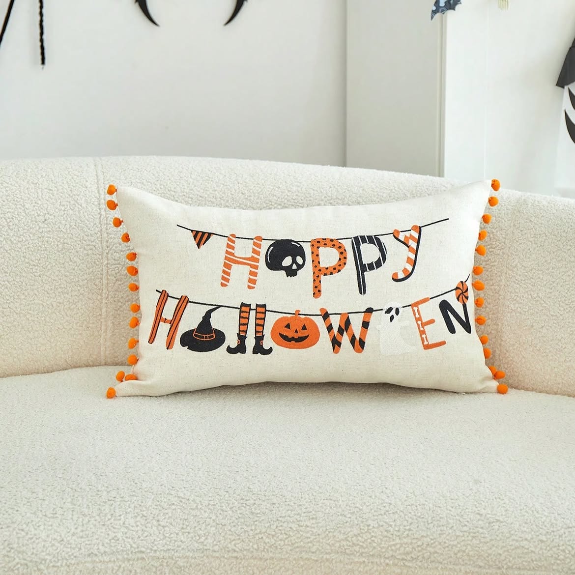 Happy Halloween embroidered lumbar cushion cover with orange pom-pom trim on cream boucle sofa with Halloween bat decorations in background