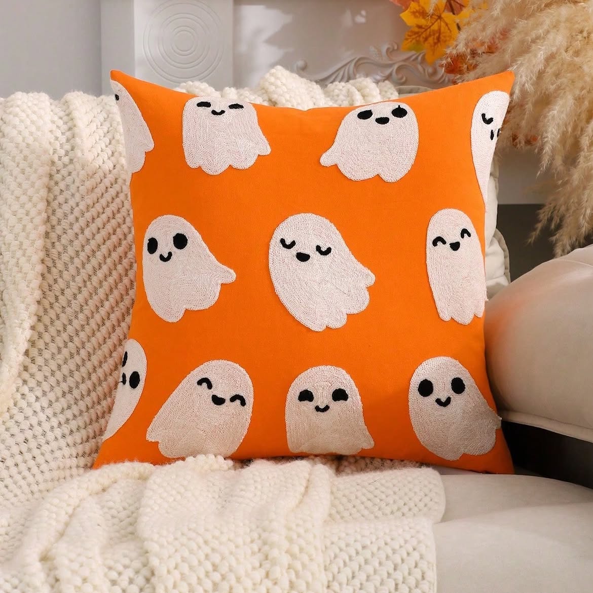 Cute ghost embroidered cushion cover in burnt orange with all-over white ghost pattern in varied expressions on cream sofa with pampas grass and autumn foliage