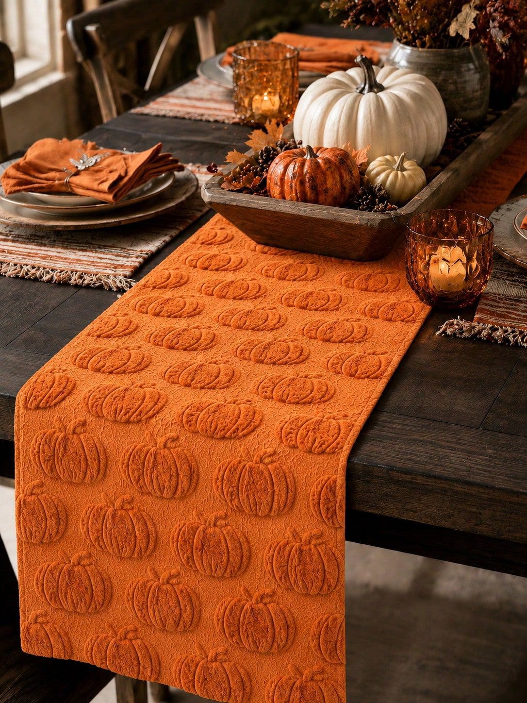 Tonal orange jacquard pumpkin table runner on dark wood dining table styled with white and orange pumpkins, candles, and autumn leaves