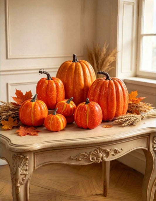 Faux orange pumpkins in assorted sizes styled on ornate cream console table with wheat stems and maple leaves in autumn setting